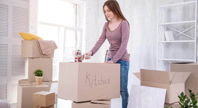 Ready to move. Pretty dark-haired girl taping the box with kitchen cutlery while packing her belongings before moving out of the apartment