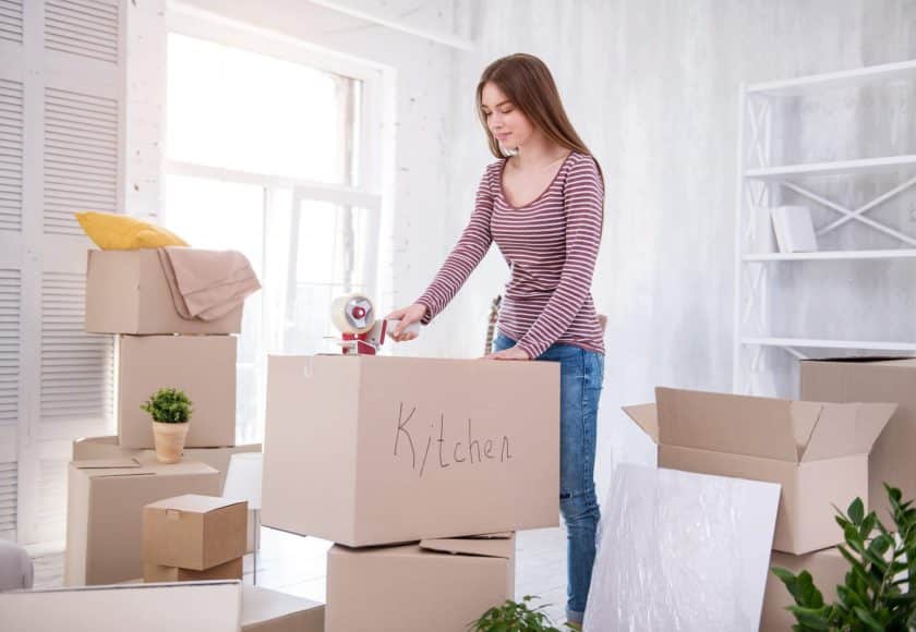 Ready to move. Pretty dark-haired girl taping the box with kitchen cutlery while packing her belongings before moving out of the apartment